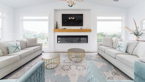 A modern living room with a fireplace, two sofas, a coffee table, and a chandelier. - Beautiful 6 bedroom beach house with pool and elevator! Steps from the beach! (Oak Island)