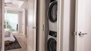 Une chambre d’hôtel avec une grande fenêtre, un lit recouvert d’une couette blanche, un lave-linge noir et blanc et un plancher en bois.