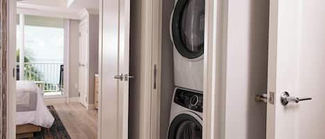 A hotel room with a large window, a bed with a white comforter, a black and white washing machine, and a wooden floor.