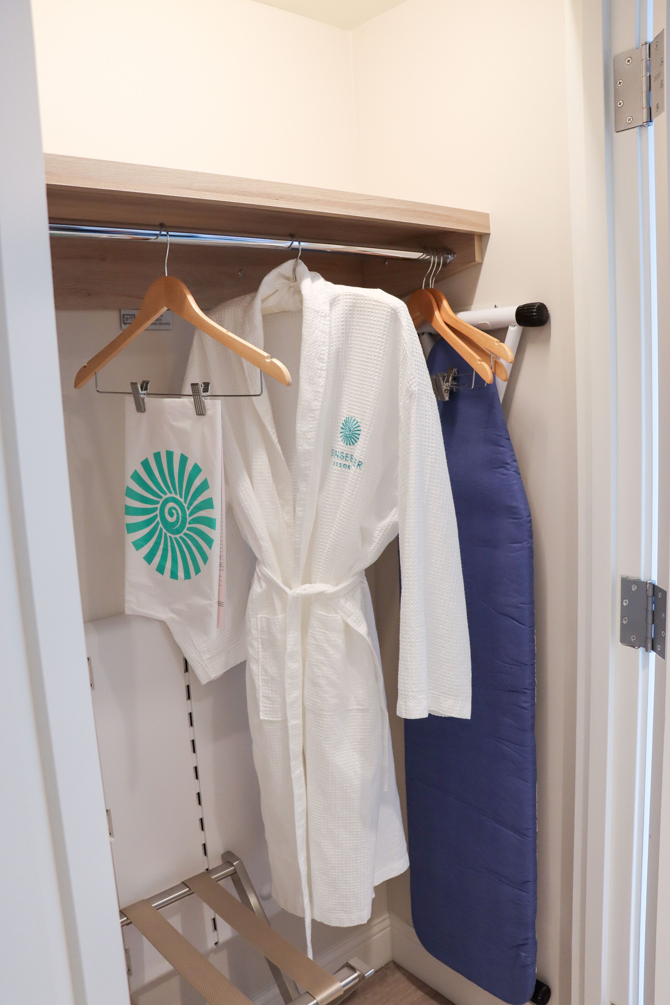 Un armario con un estante de madera, una bata blanca con un logotipo, una prenda azul y una toalla blanca con un logotipo verde.
