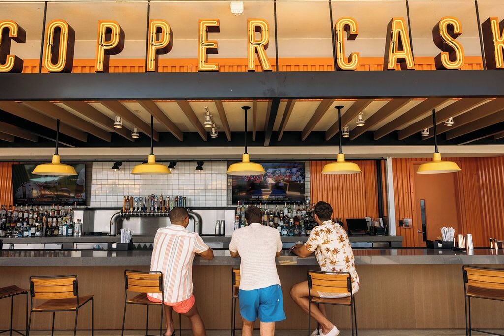 A bar area with a neon sign reading "COPPER CASINO" above the counter. There are three people at the bar, two standing and one sitting. The bar has a tiled backsplash and a variety of bottles and glasses on the shelves.