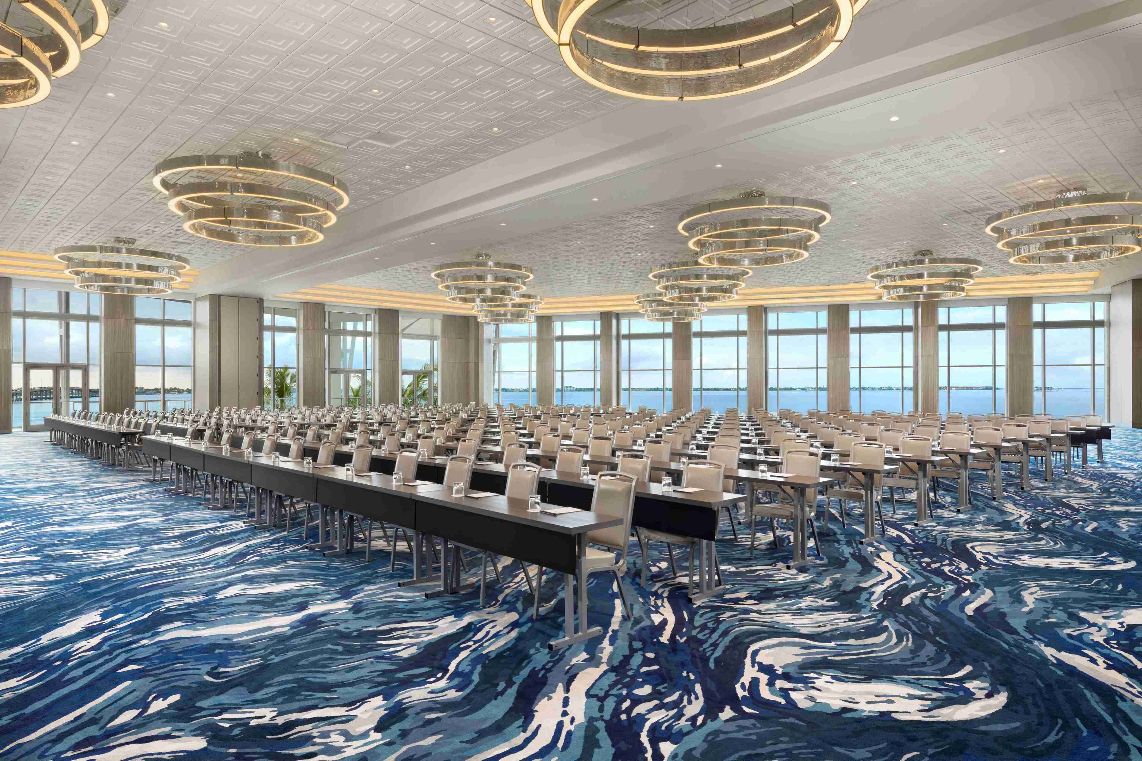 A large conference room with rows of tables and chairs, large windows, and modern circular chandeliers.