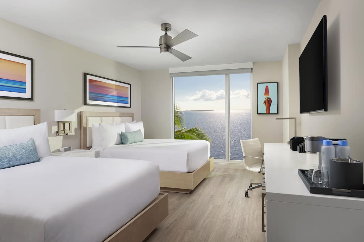Een hotelkamer met twee bedden, een bureau, een flatscreen-tv, een plafondventilator en een groot raam met uitzicht op de oceaan.