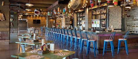 A restaurant with long communal tables and blue chairs, a bar area with shelves of bottles, and hanging decorative lights.