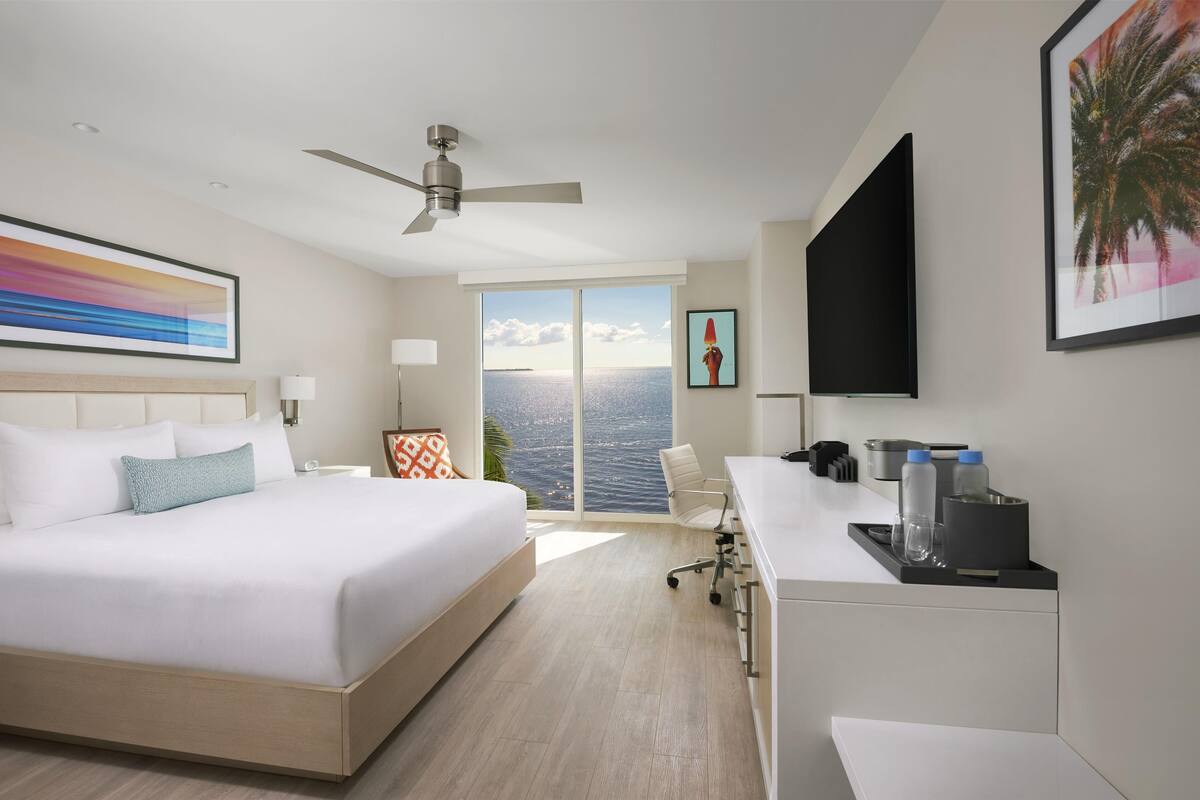 Une chambre d’hôtel équipée d’un lit, d’un bureau, d’une chaise, d’un ventilateur de plafond, d’une télévision et d’une grande fenêtre donnant sur un beau paysage.