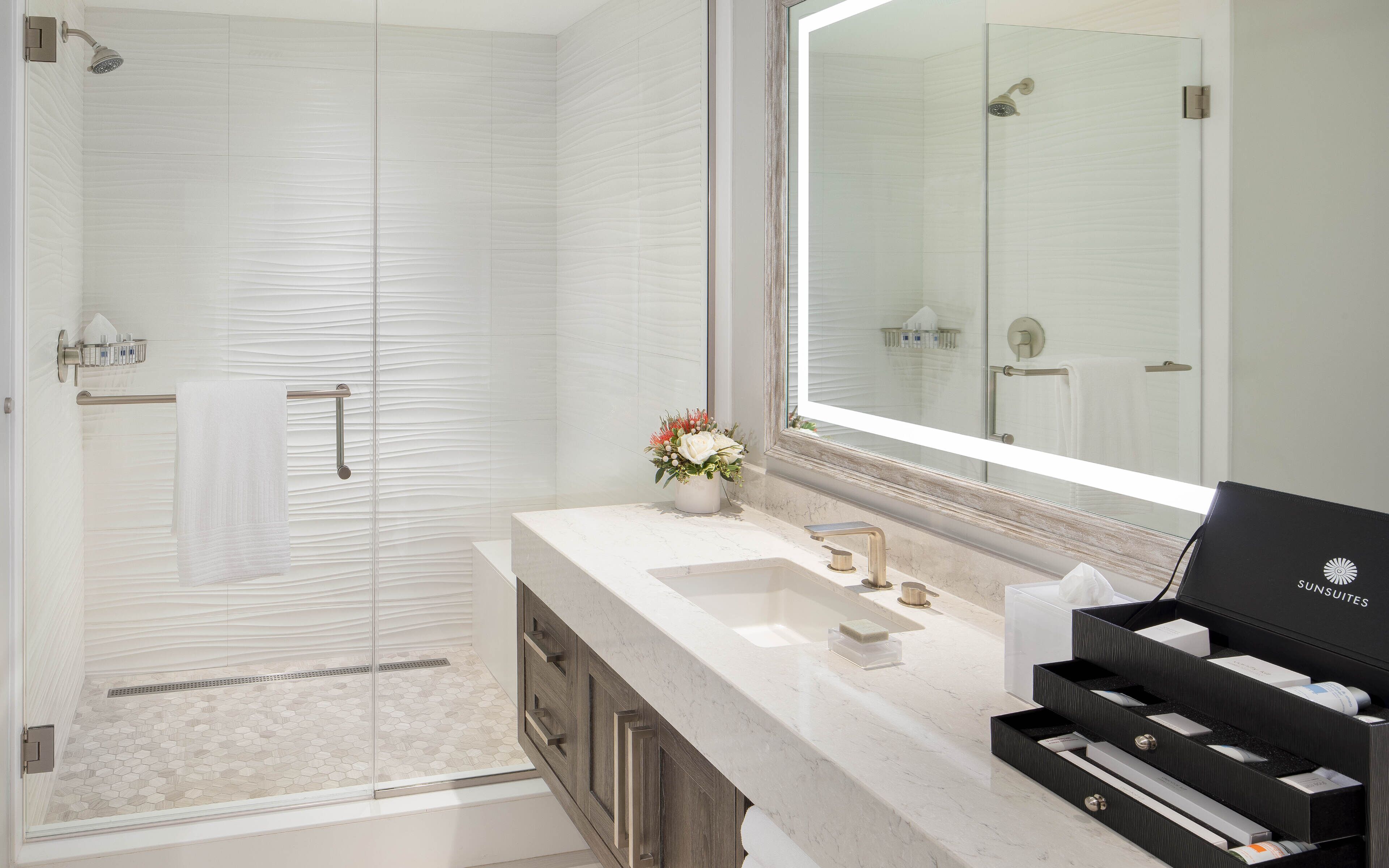 Un bagno moderno con box doccia in vetro, un vanità con uno specchio grande e un mobile contenitore nero etichettato 'SUNSUITES'.
