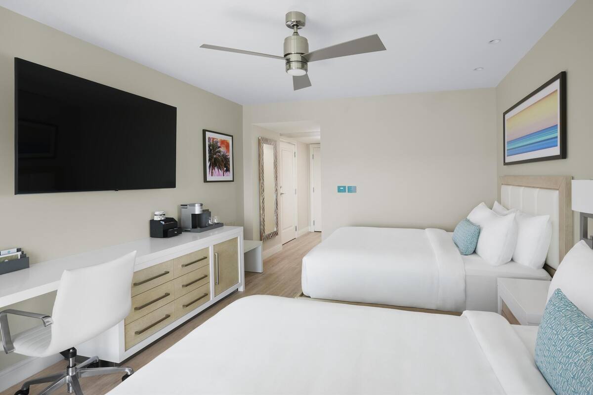 Une chambre d’hôtel comprenant un lit, un bureau avec une chaise, une télévision, un ventilateur de plafond et un tableau au mur.