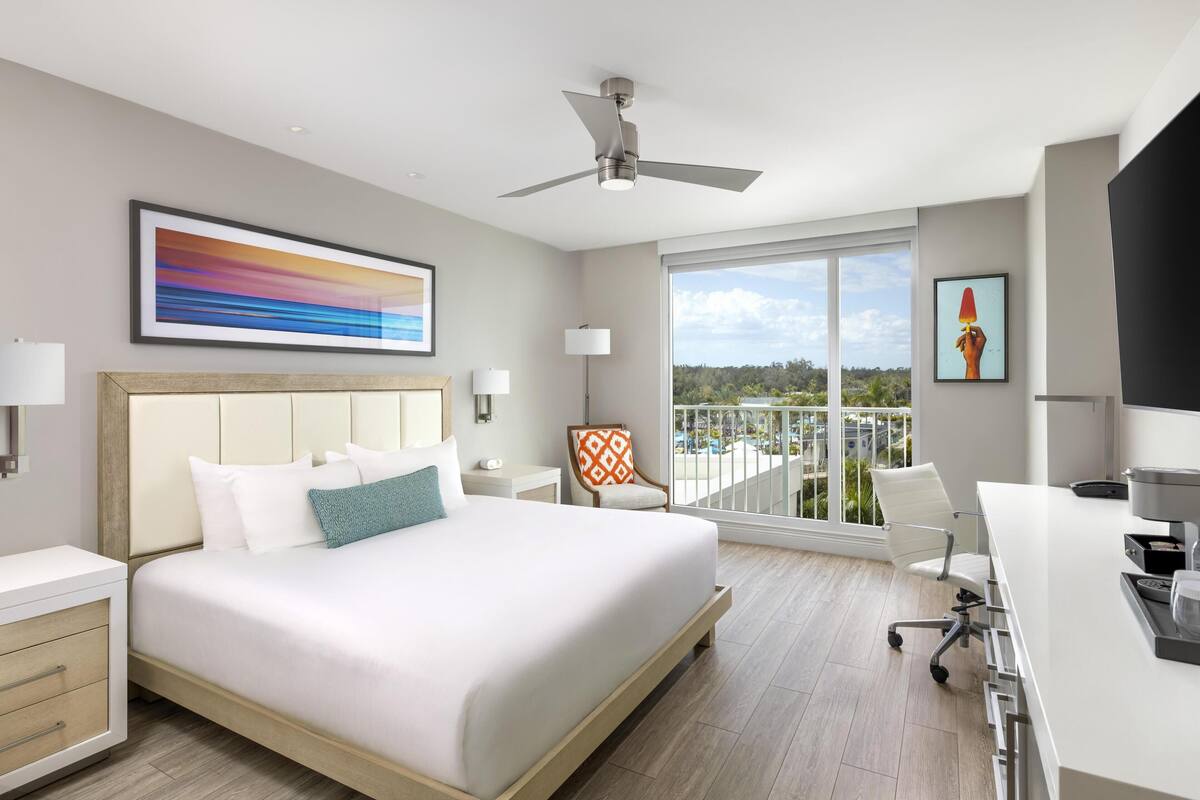 Une chambre d’hôtel équipée d’un lit, d’un bureau, d’une chaise, d’un ventilateur de plafond, d’une grande fenêtre et d’œuvres d’art accrochées aux murs.