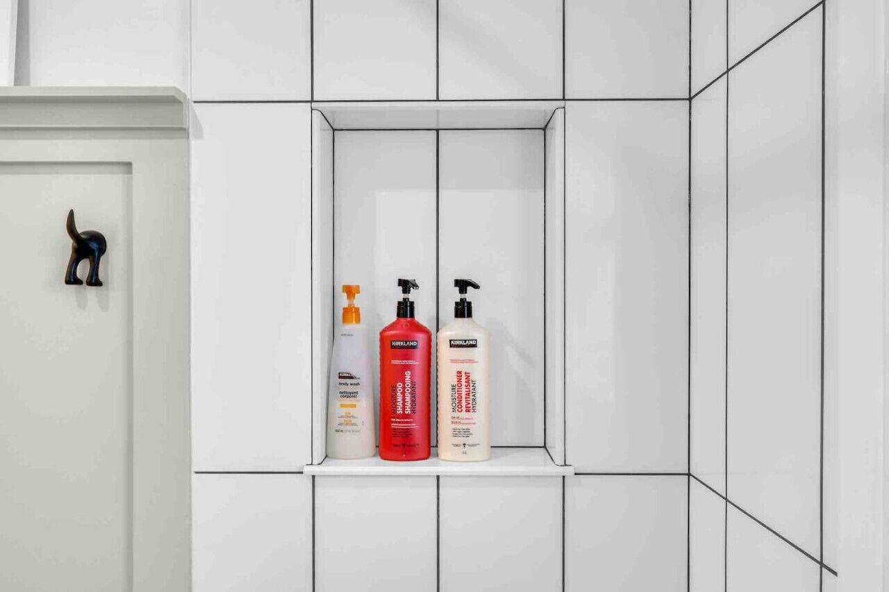 A bathroom with white tiled walls, a white cabinet, and three bottles of Kirkland body wash, shampoo, and conditioner.