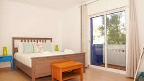 Une chambre avec un lit en bois, un petit banc, une table de chevet et une grande porte-fenêtre coulissante donnant sur un balcon.