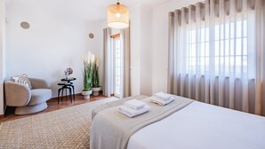 A hotel room with a large bed, a sitting area with a chair and a small table, a potted plant, and a view through sheer curtains.
