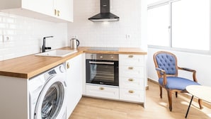 A compact kitchen with a washing machine, oven, and a chair near a window. - SERAFIM BEACH APARTMENT - Heart of the Old Town (Albufeira)