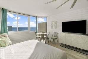 Un dormitorio con cama, televisión, mesa de comedor con sillas y vista al océano.