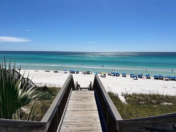 A wooden boardwalk leading to a beach with umbrellas and people.