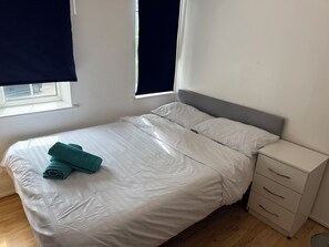 A neatly made bed with white bedding and a teal towel, a bedside drawer unit, and a window with a view of the outside.