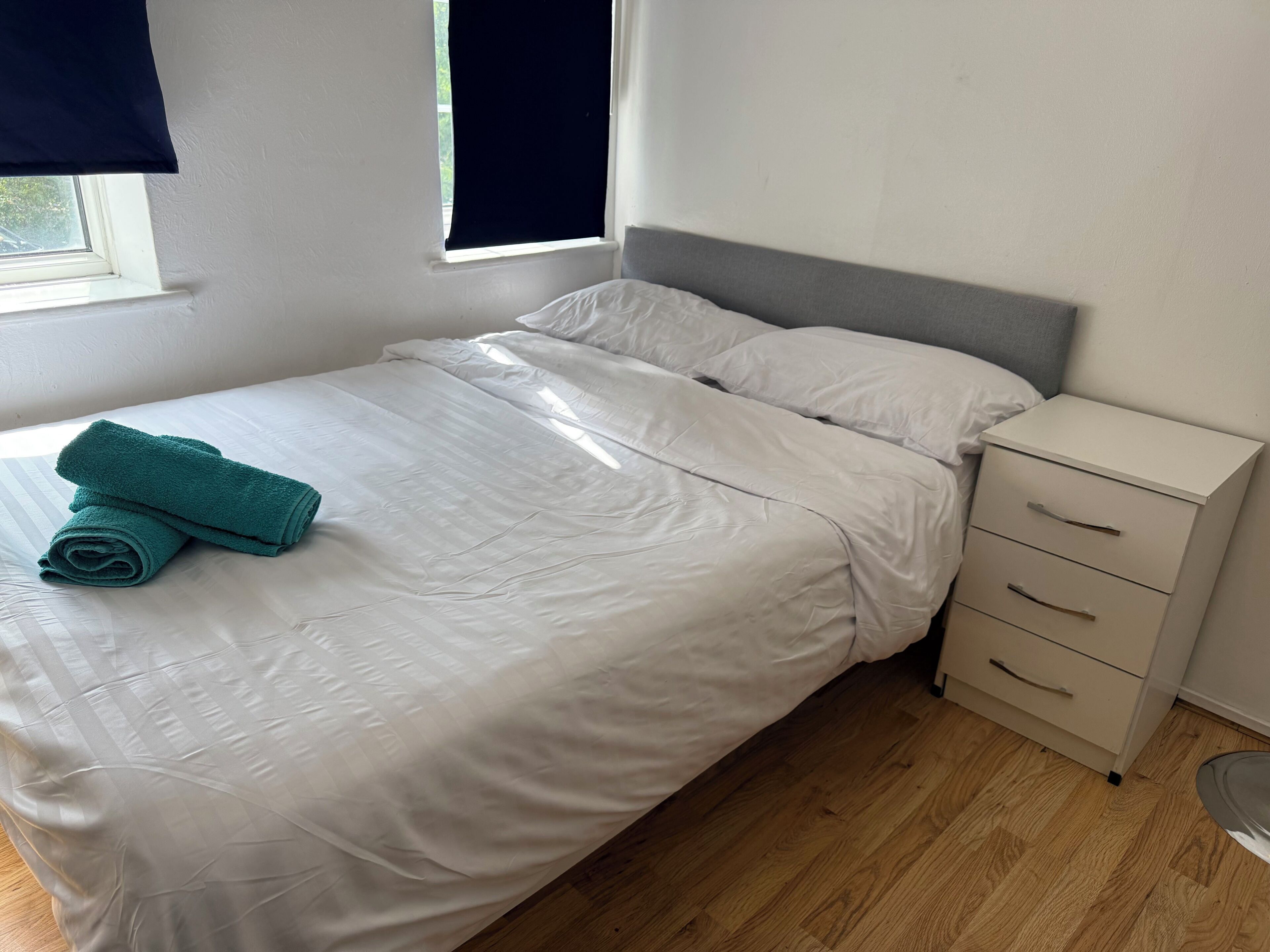 A neatly made bed with white bedding and a teal towel, a bedside drawer unit, and a window with a dark blind.