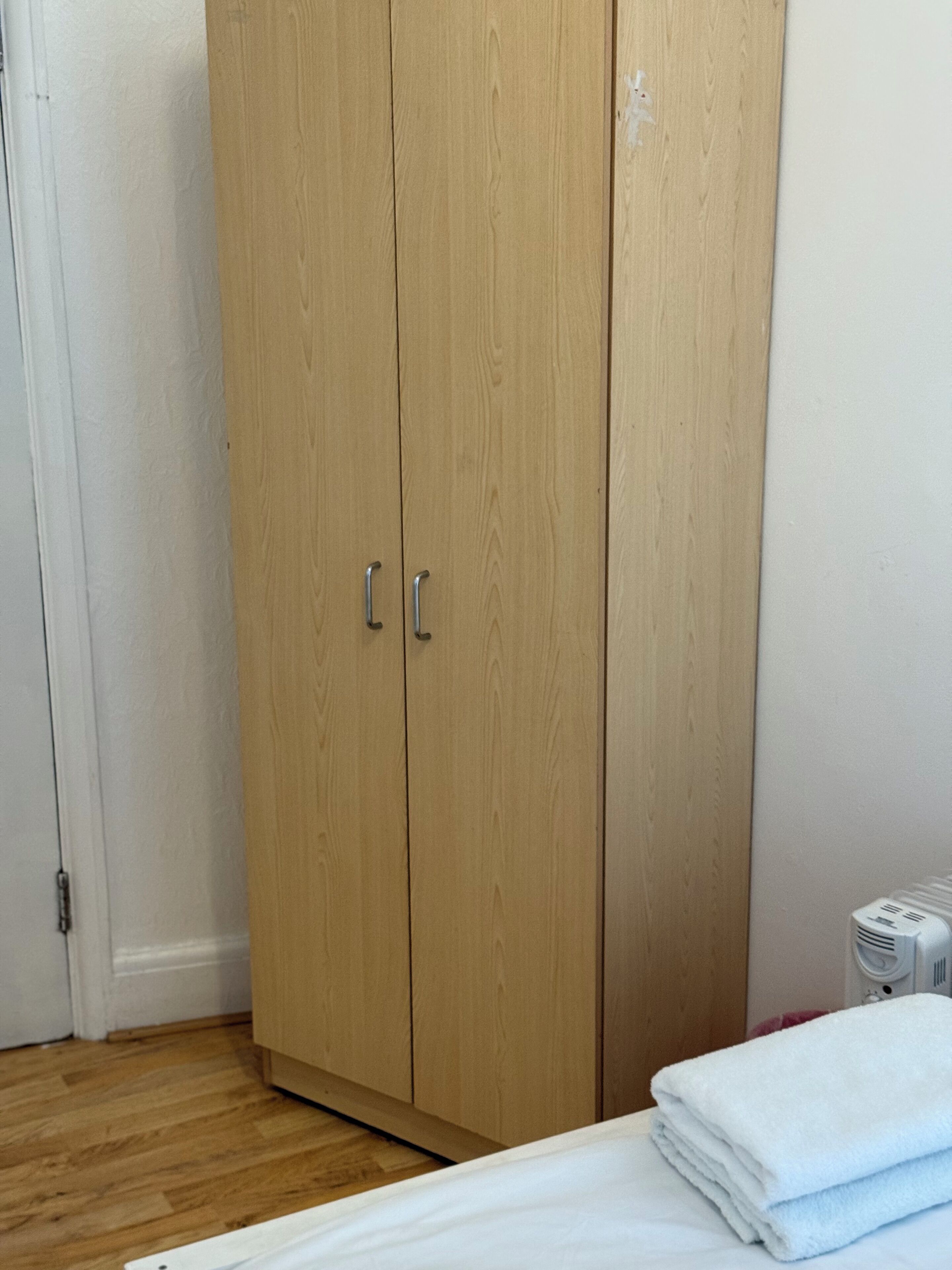 Een slaapkamer met een houten garderobe, een radiator, en een keurig opgevouwen witte handdoek op het bed.