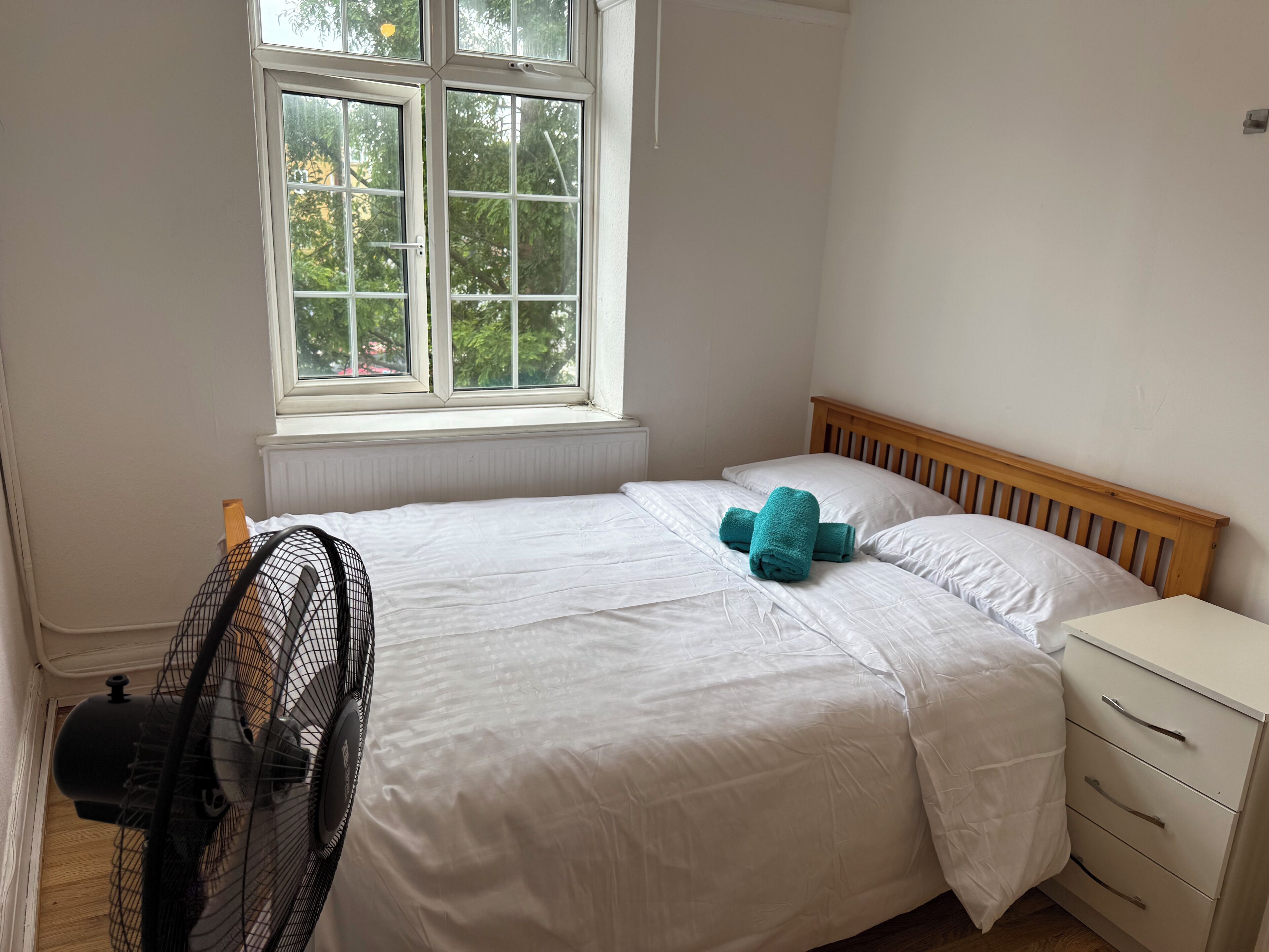 Een slaapkamer met een bed, een houten hoofdbord, een nachtkastje en een raam met uitzicht op bomen.