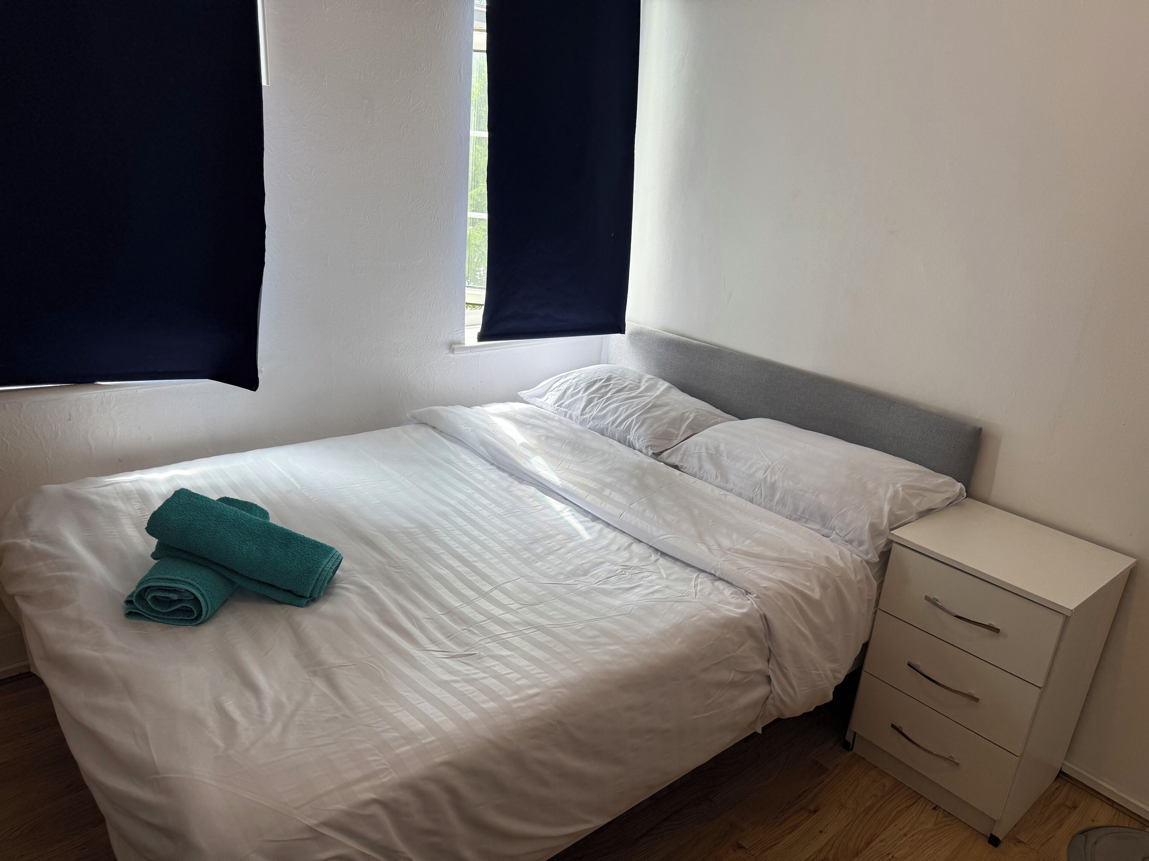 Una cama bien hecha con sábanas blancas y una toalla color turquesa, un mueble cajonera junto a la cama y una ventana con cortinas oscuras.