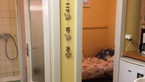 A small, compact room with a shower, a bed with a floral bedspread, and a wooden table with a woven placemat. - Le Petit Brit (Syens)