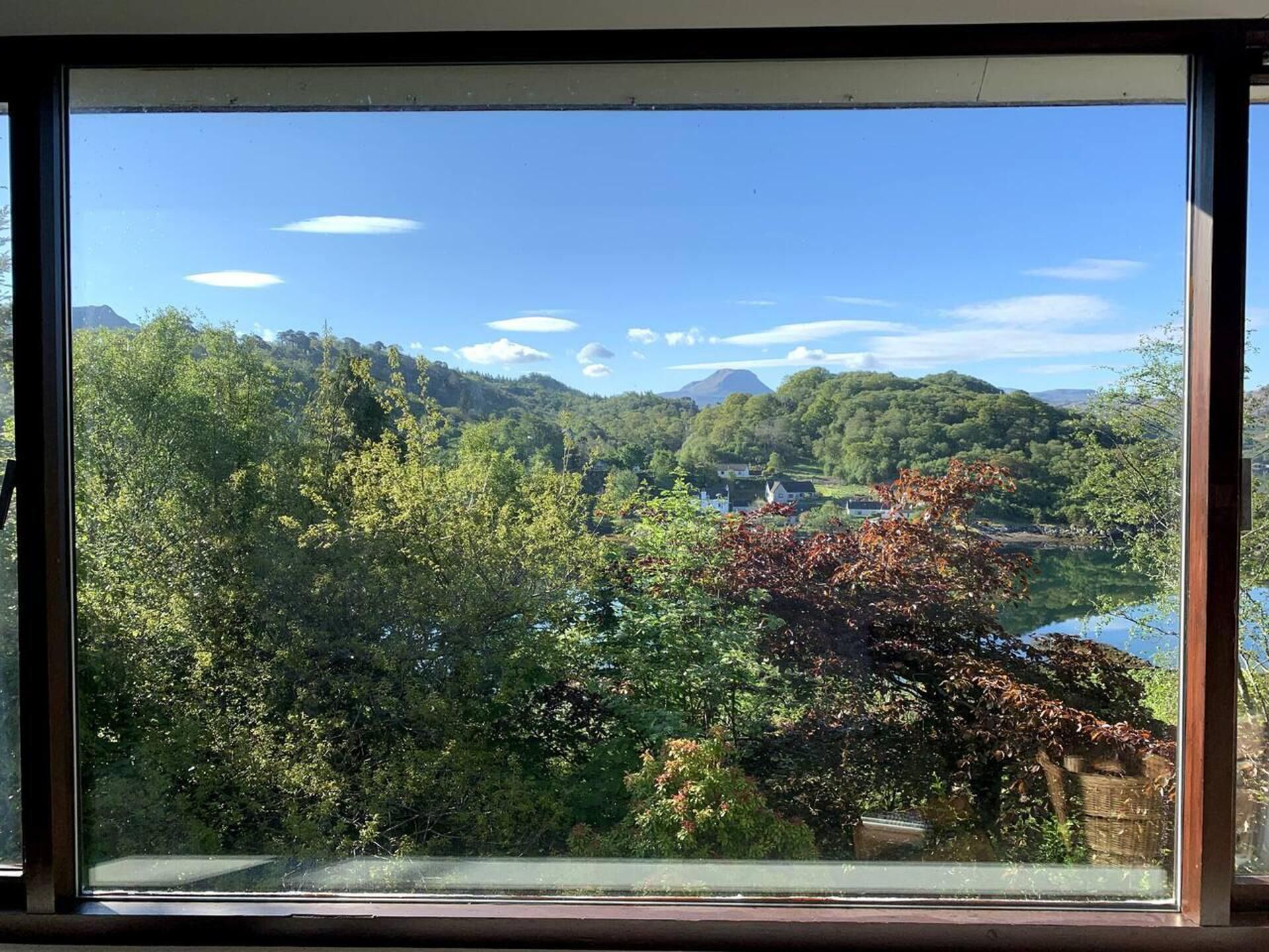 Une vue à travers une fenêtre sur un paysage verdoyant et luxuriant, avec une montagne à l’horizon et un ciel bleu dégagé.