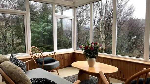 A conservatory with a wooden bench, a round table, and a vase of flowers. - Midcentury 3-bed House in Gairloch (Gairloch)