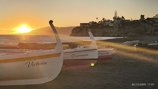 Barcos chamados Victoria, Boquerona e Margarita numa praia com o pôr do sol ao fundo.