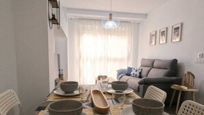 Uma área de refeições com uma mesa posta para quatro pessoas, incluindo tigelas, pratos e talheres. Há um sofá e uma pequena mesa lateral ao fundo.