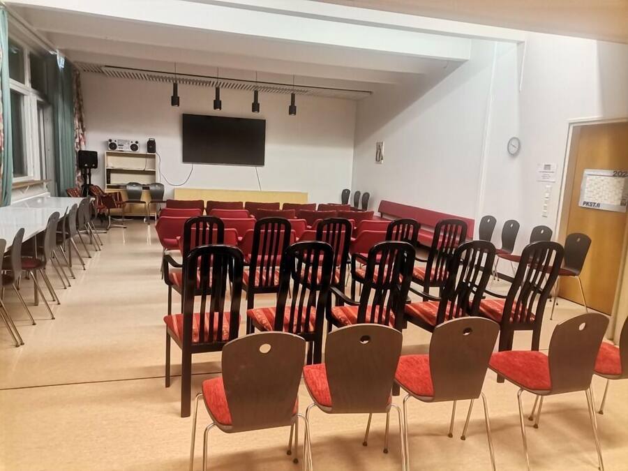 Un salón de conferencias con filas de sillas rojas y una mesa larga, un televisor de pantalla plana montado en la pared y un reloj en la pared.