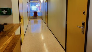 Un pasillo con puertas numeradas 6, 7 y 8, que conducen a habitaciones. Un escritorio de madera con una silla está a la izquierda, y se observa un botiquín de emergencia verde en la pared.
