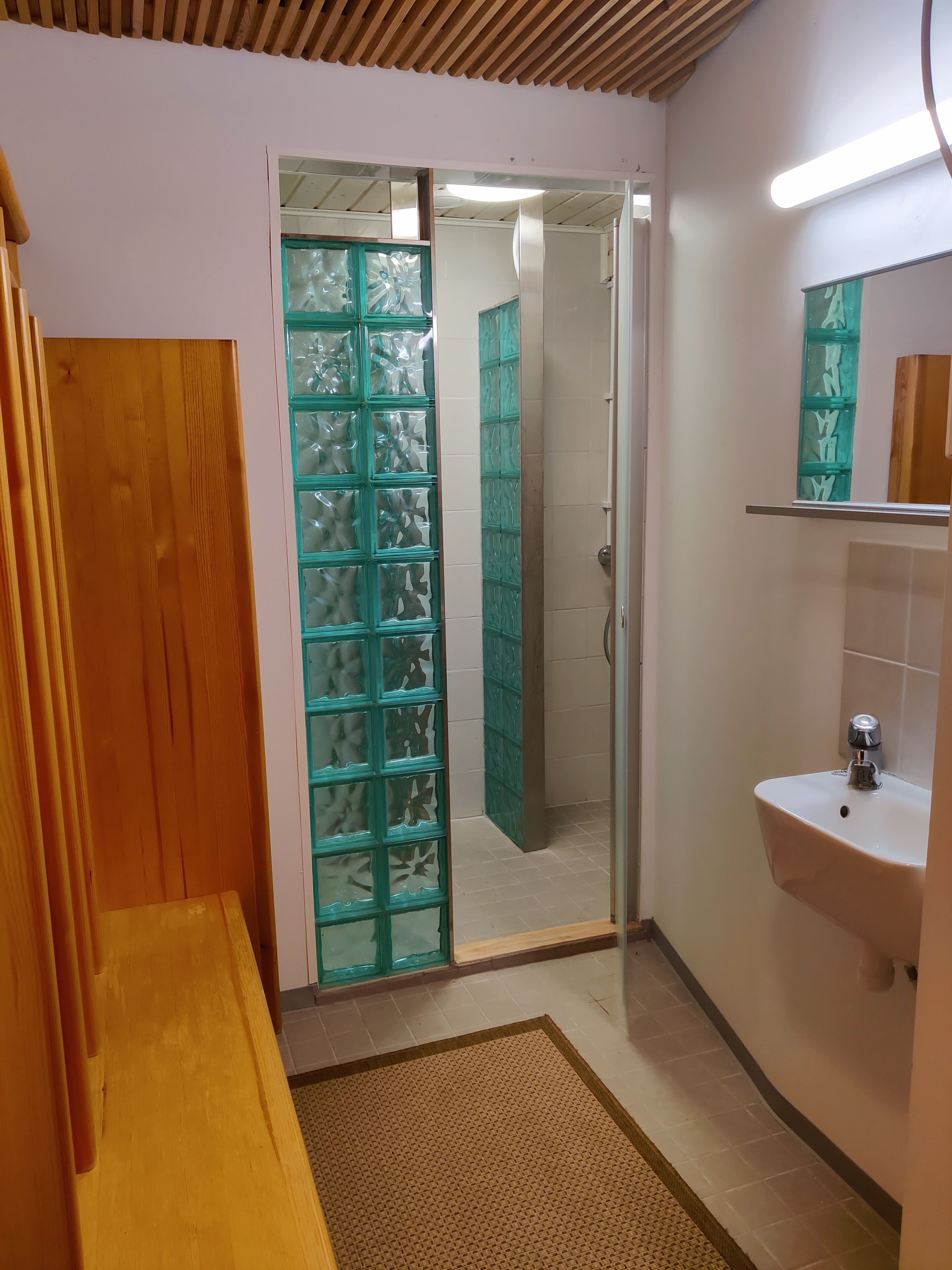 Un baño con banca de madera, lavamanos y puerta de vidrio que da a la zona de la ducha.