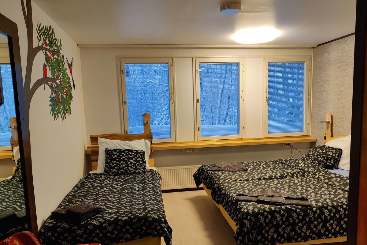 A room with two single beds, a wooden headboard, a window with blinds, a radiator, and a wall-mounted shelf.