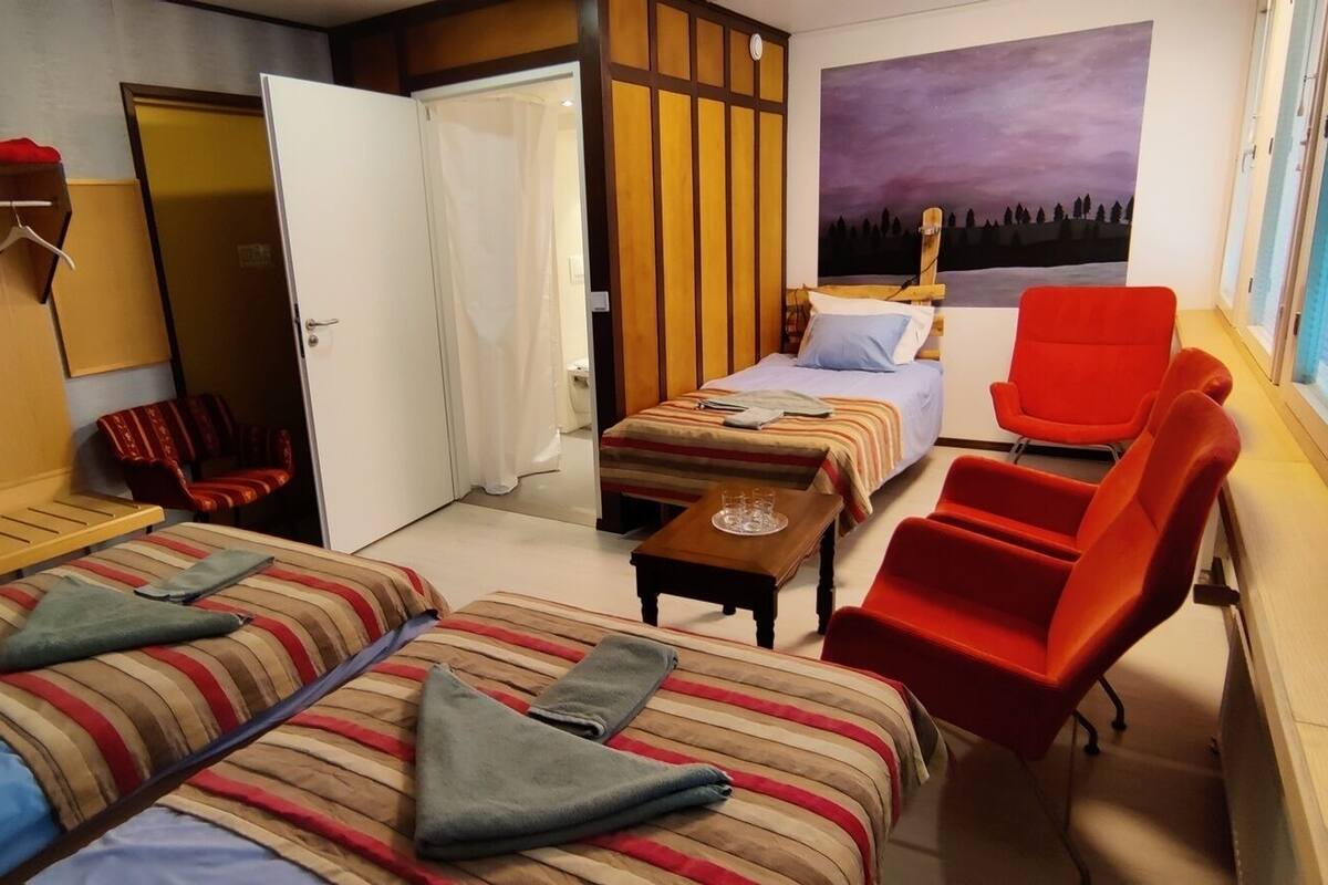A hotel room with two beds, a red armchair, a small table, and a wardrobe.