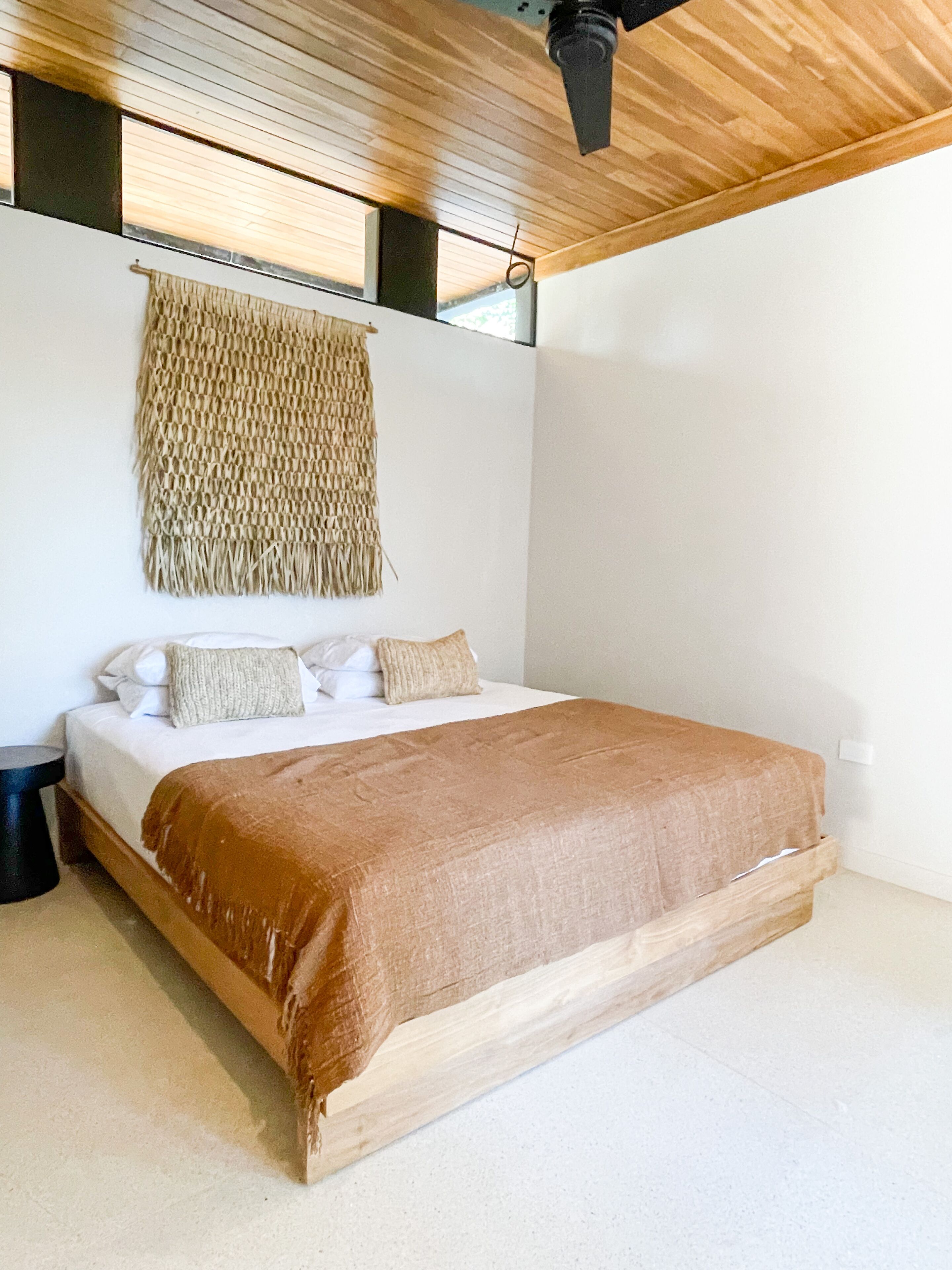 A bedroom with a wooden bed frame, white bedding, and a woven wall hanging.