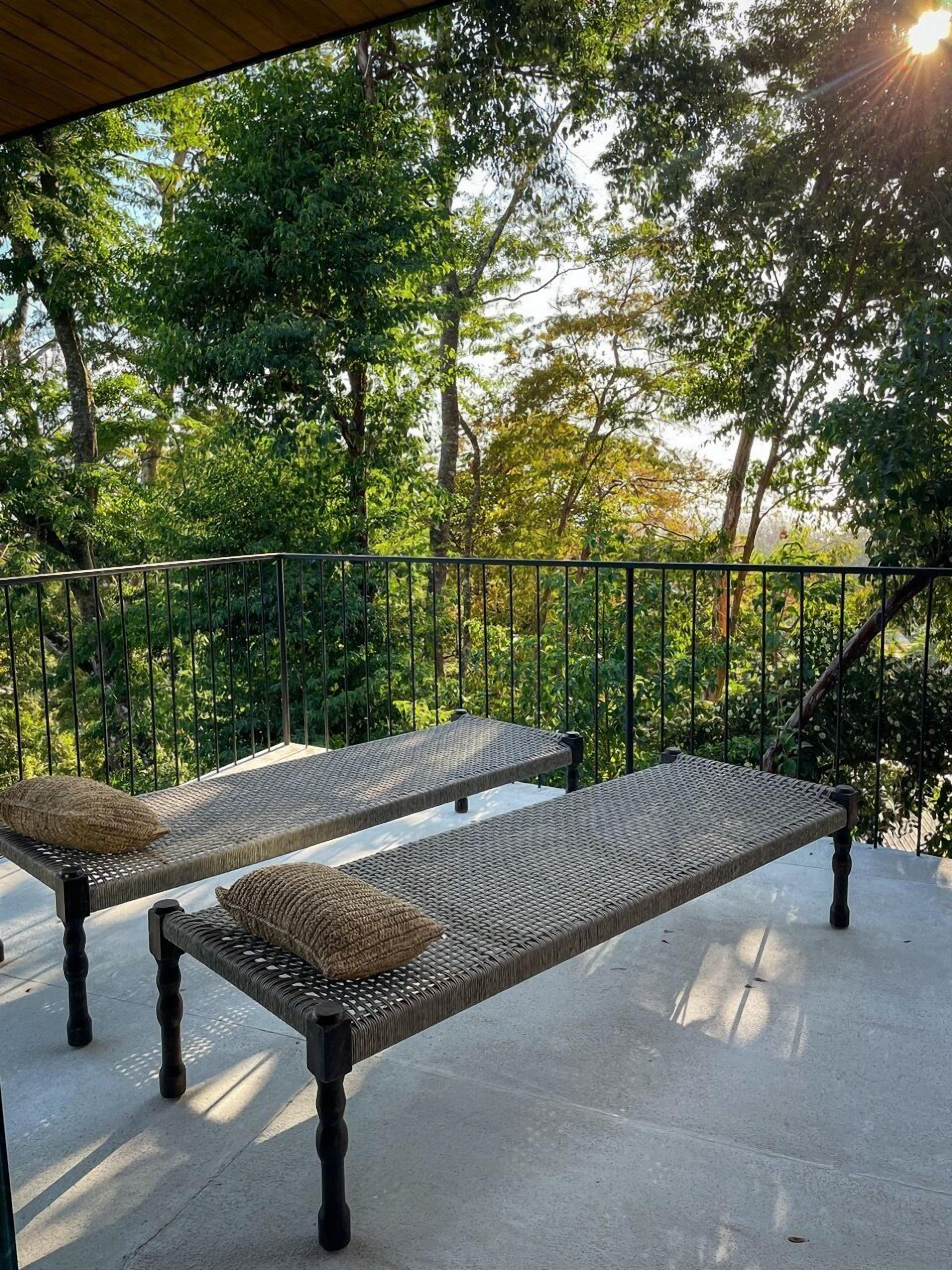 Two wicker lounge chairs with cushions on a balcony with a metal railing, overlooking a dense green forest.
