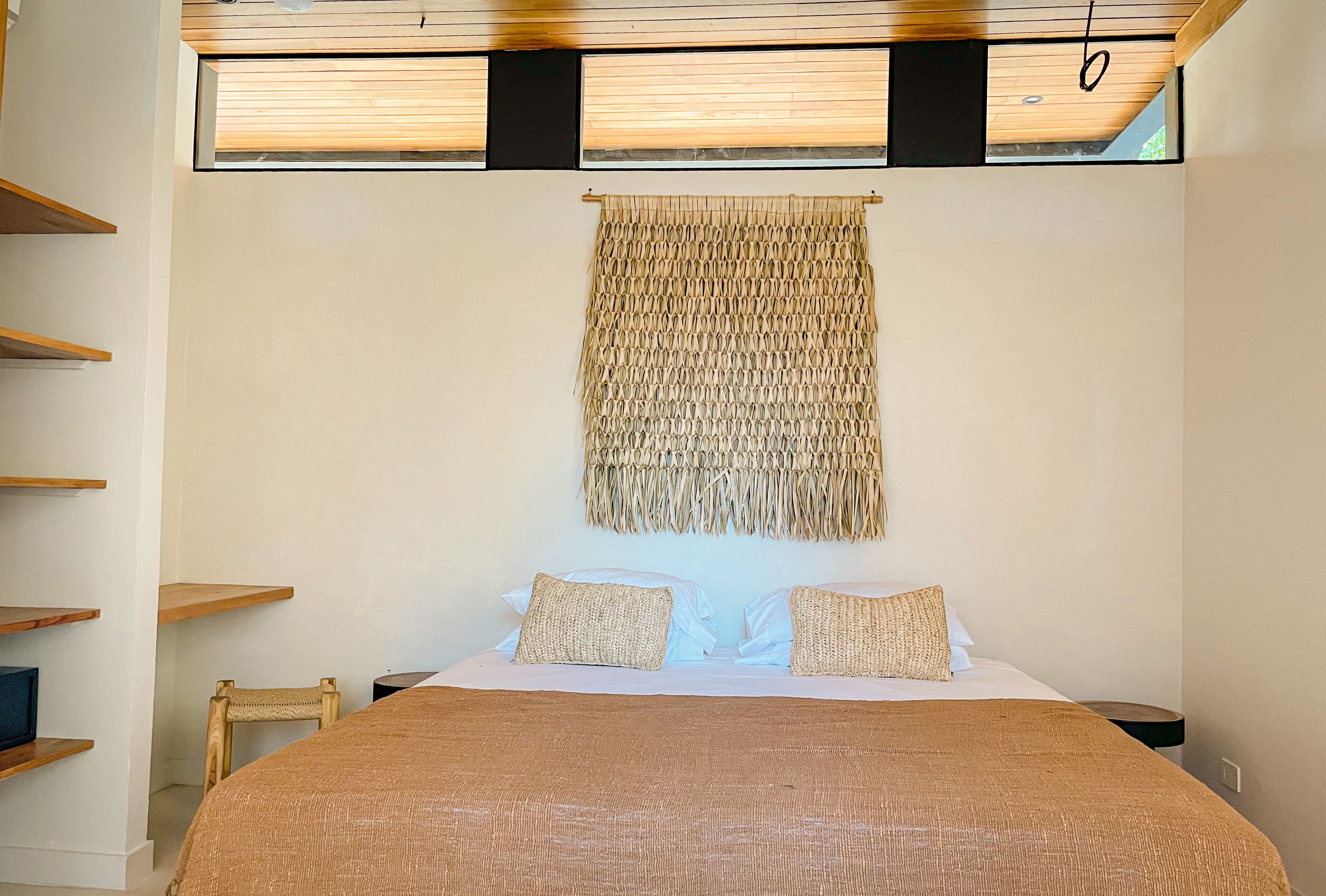 A bedroom with a large bed, two pillows, a woven tapestry, and wooden shelves.