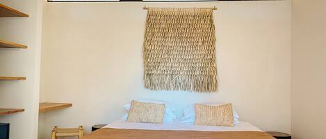A bedroom with a large bed, two pillows, a woven tapestry, and wooden shelves.