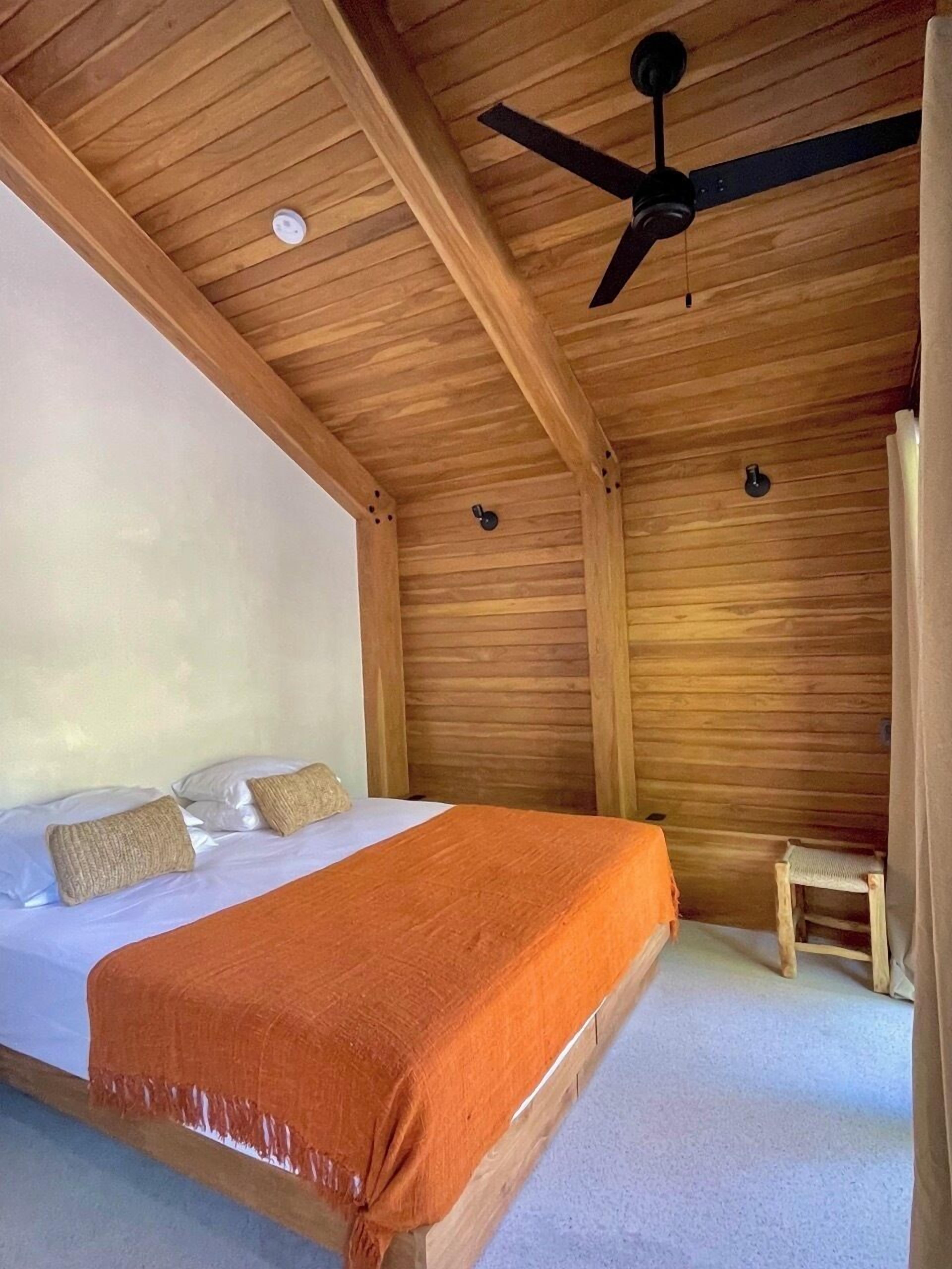 Ein Zimmer mit Holzwänden, einem Bett, einem Deckenventilator und einem kleinen Holzschemel.