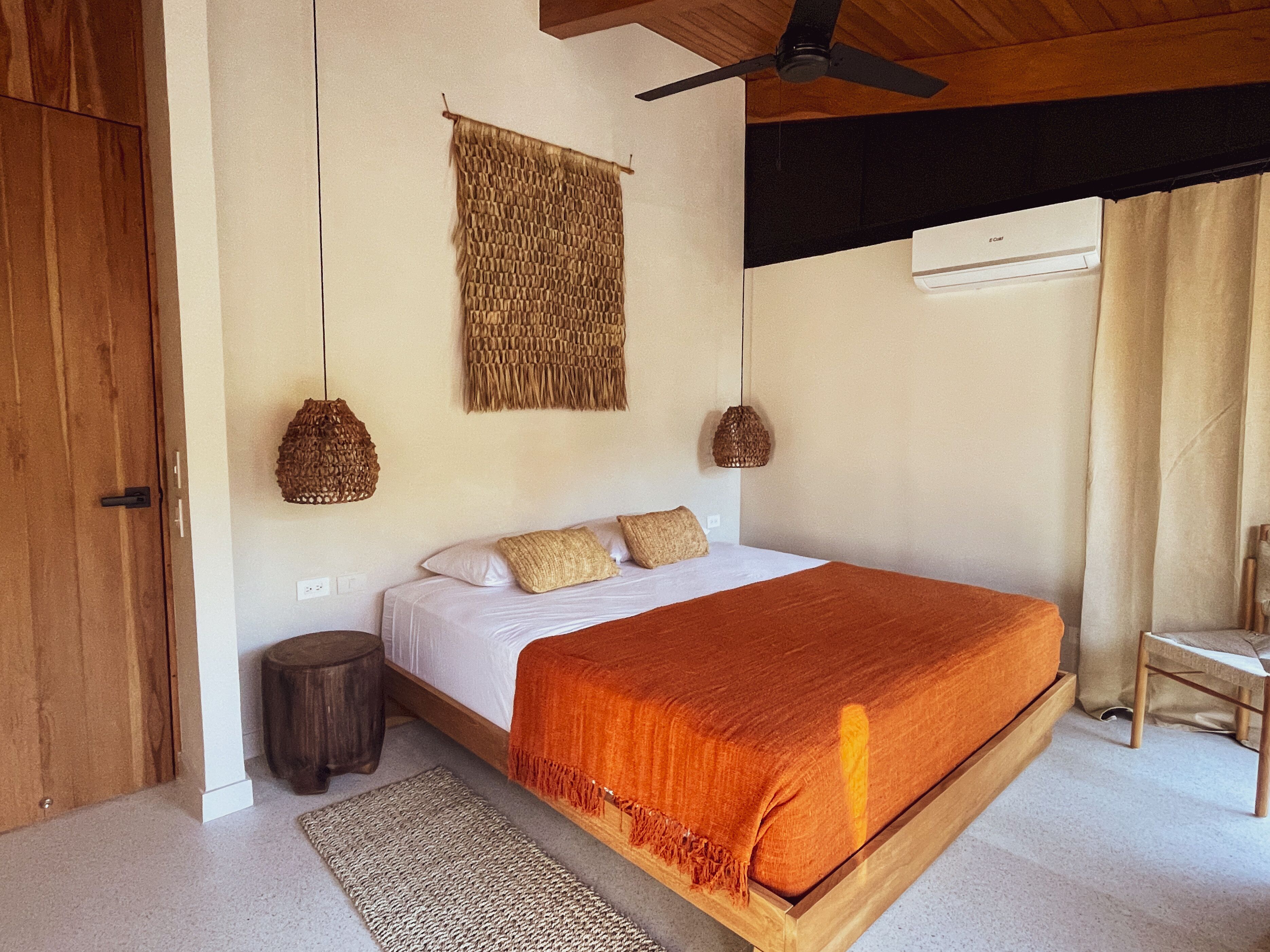 A bedroom with a wooden bed, a ceiling fan, woven wall hangings, and a small round stool.