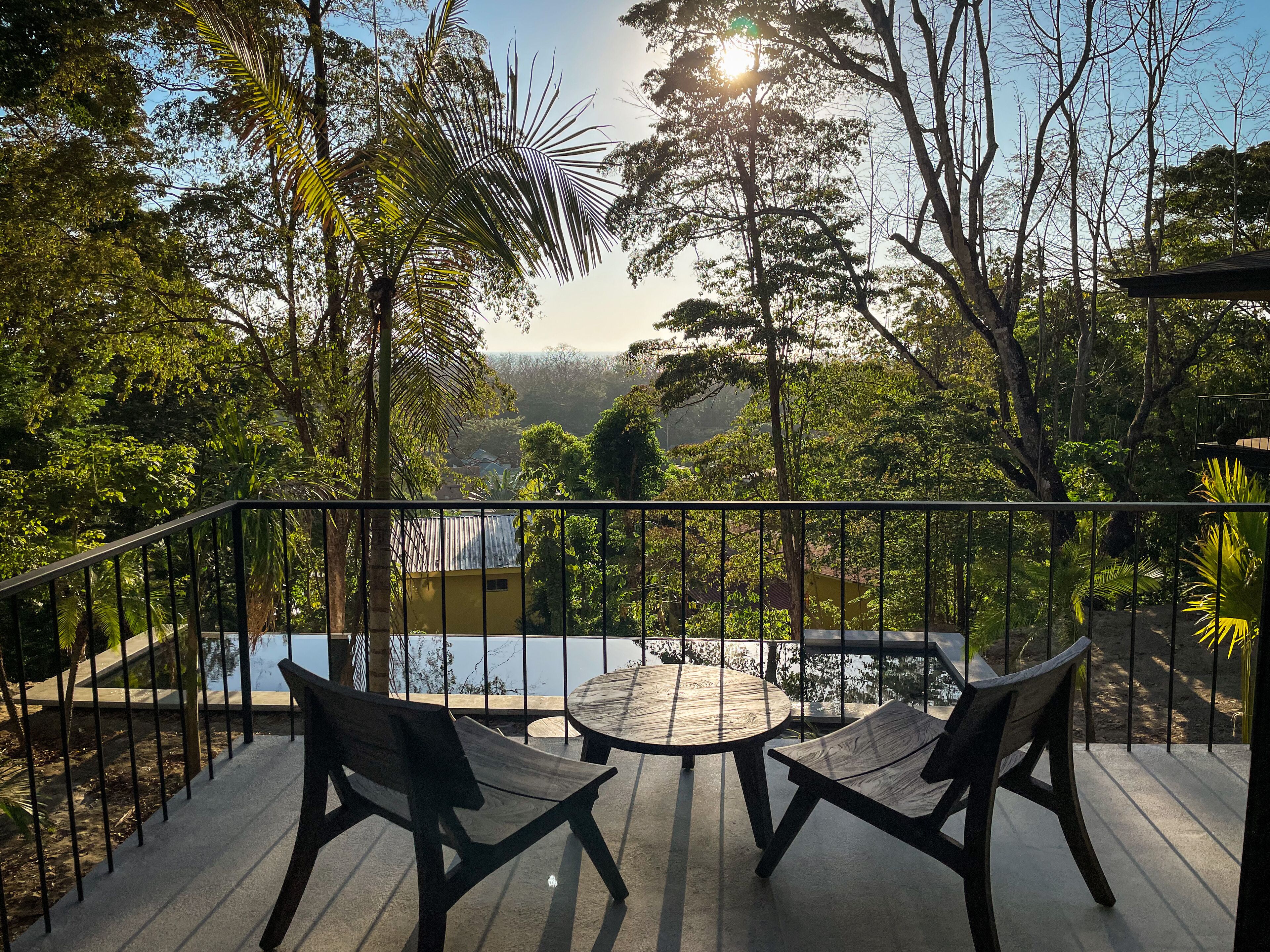 A deck with a round table and two chairs, surrounded by a metal railing, overlooking a pool and lush greenery.