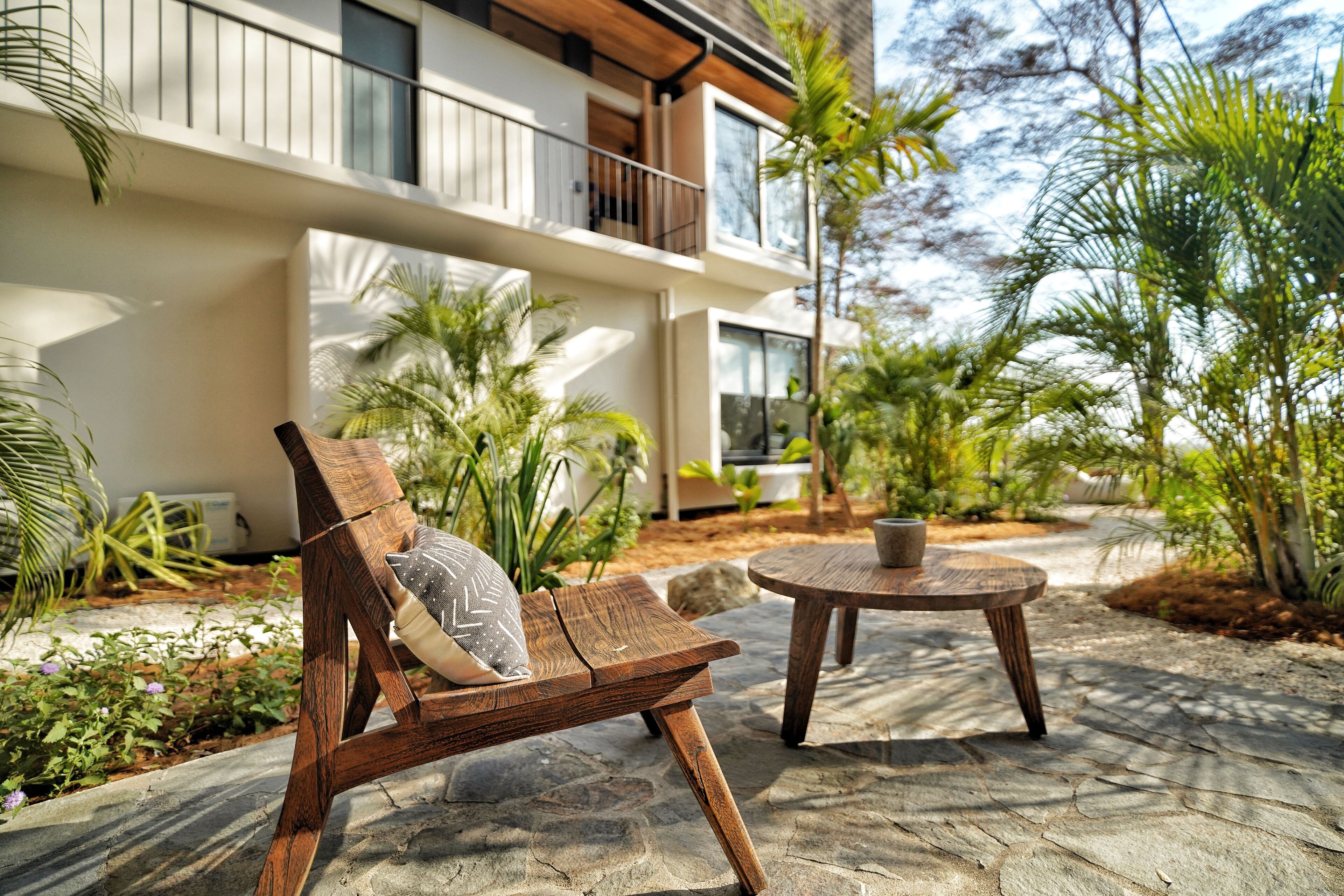 Un fauteuil de jardin en bois avec un coussin, une petite table ronde et une terrasse en pierre agrémentée de plantes, avec un bâtiment visible au loin.