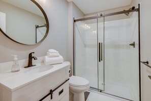 Un baño moderno con ducha envuelta en vidrio, espejo redondo, lavamanos blanco y un inodoro.
