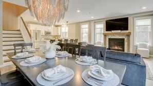 A modern dining area with a long table set for six, a fireplace, and a staircase leading to the upper floor.