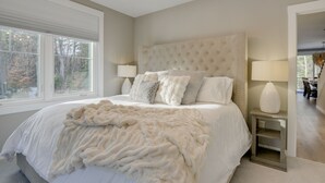A bedroom with a large bed, two bedside lamps, a tufted headboard, and a view of trees through the window.