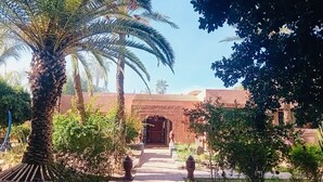 A courtyard with a palm tree, a pathway leading to a building with a traditional archway, and a clear blue sky. - Villa Avec Piscine Privée au Cœur de la Palmeraie (Marrakech)