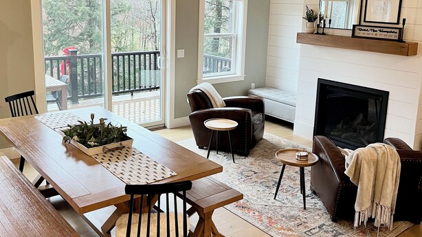A cozy living room with a wooden dining table, a fireplace, and a large window overlooking a balcony.