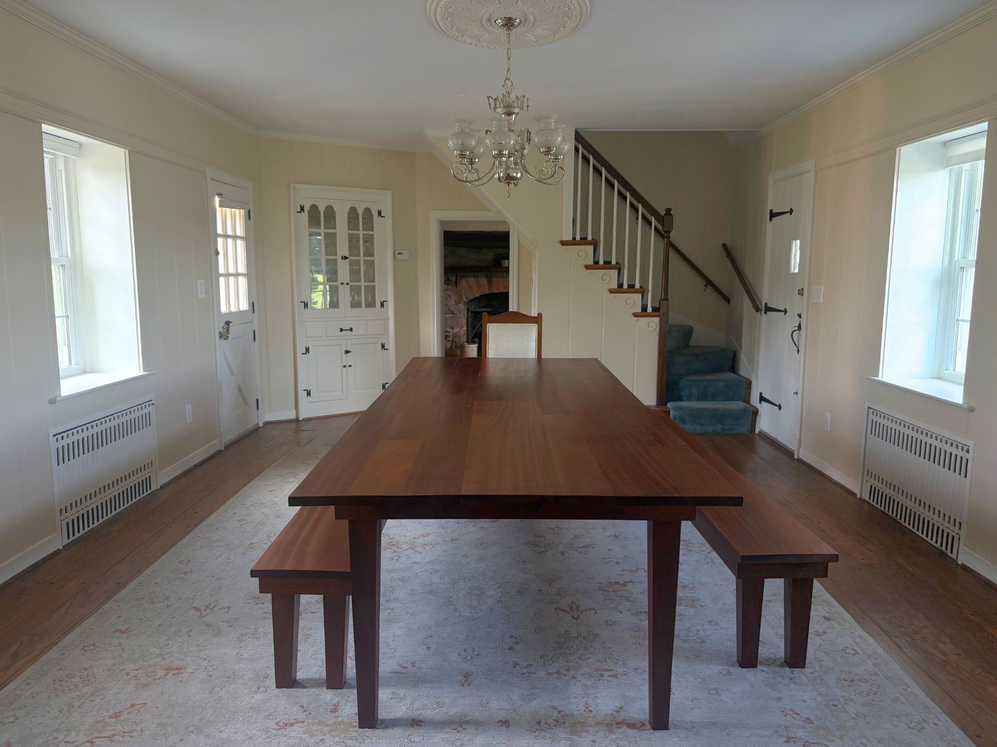 A spacious dining room with a long wooden table and benches, a chandelier, and a staircase leading to an upper level.