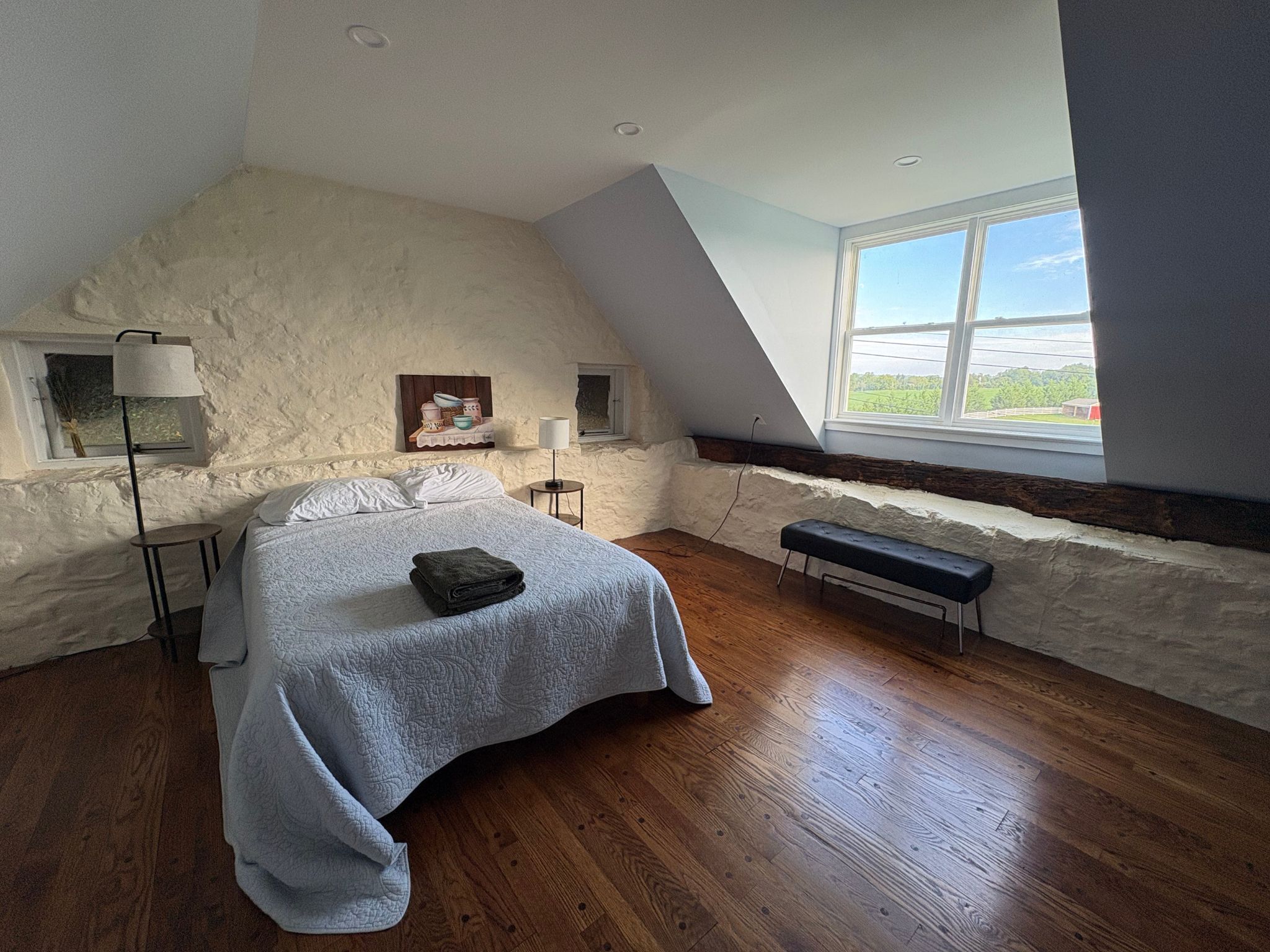 A bedroom with a sloped ceiling, a bed with a quilt, a bench, a small table, a lamp, and a painting on the wall.