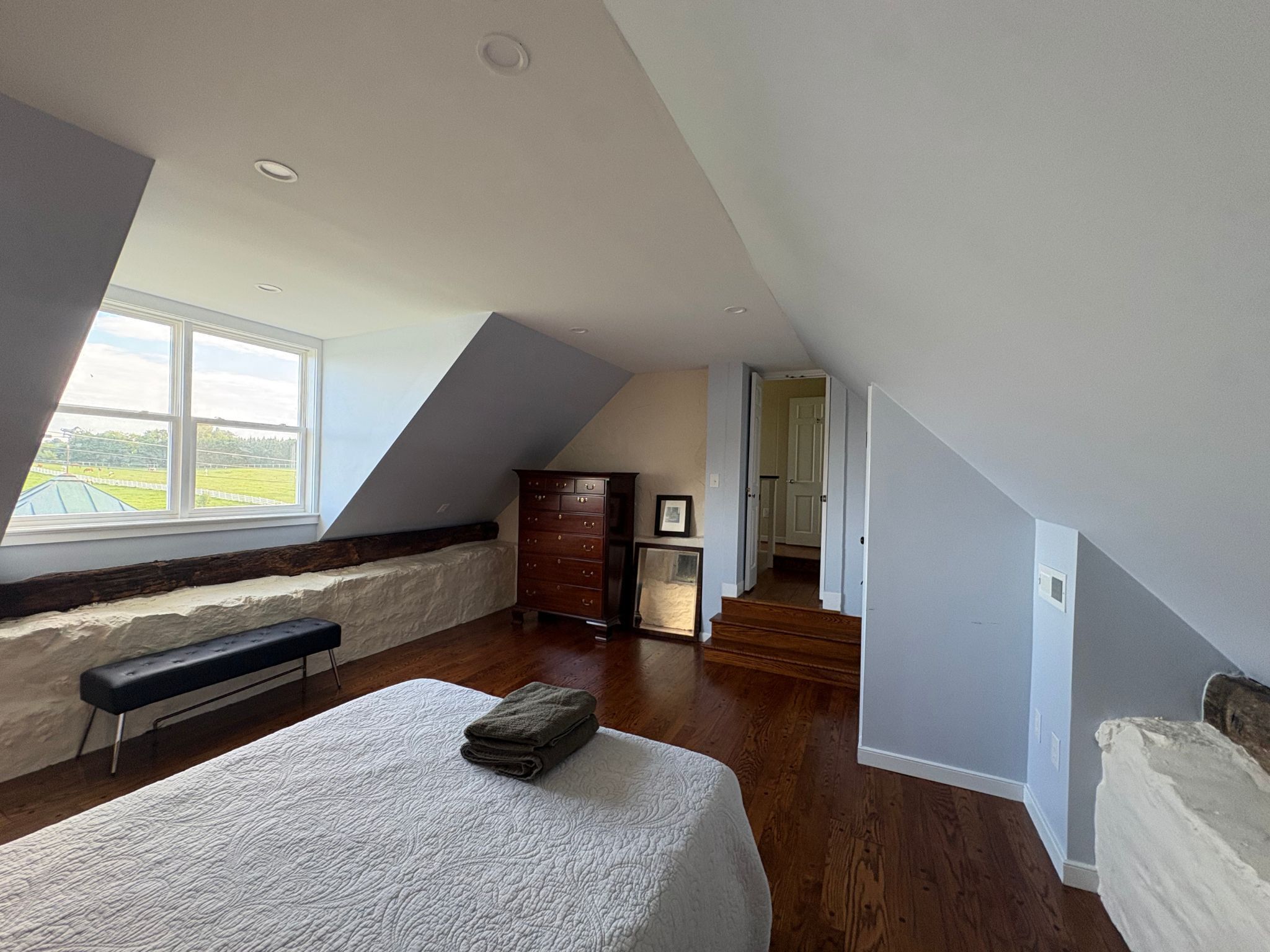 A bedroom with a sloped ceiling, a large window, a bed, a dresser, and a bench.