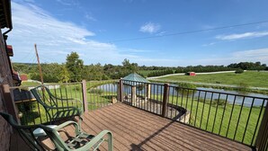 A wooden deck with metal railings, two green chairs, and a view of a green field and a small body of water. - Peaceful Retreat in the Countryside with Farm Animals and Play Area (Boyds)